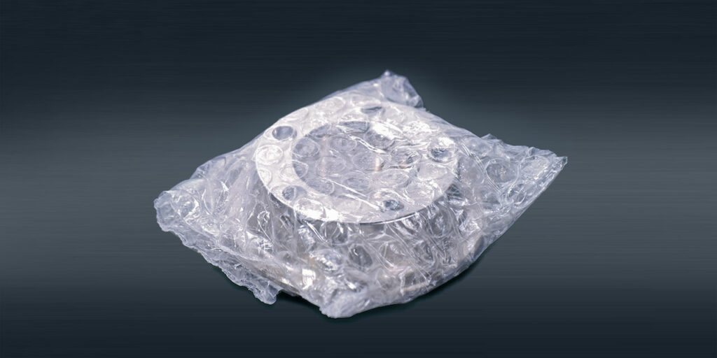 Rundes Metallteil mit Bohrungen, in einem Beutel aus Luftpolsterfolie verpackt